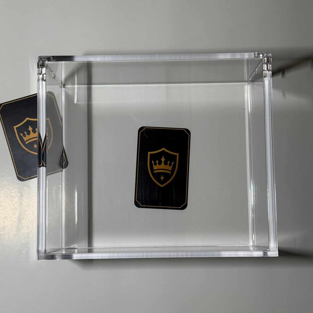 A clear acrylic display box on a light gray background, containing a small black shield-shaped card with a gold crown logo. A second matching card is partially visible on the left side.