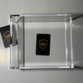 A clear acrylic display box on a light gray background, containing a small black shield-shaped card with a gold crown logo. A second matching card is partially visible on the left side.