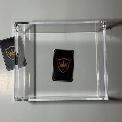 A clear acrylic display box on a light gray background, containing a small black shield-shaped card with a gold crown logo. A second matching card is partially visible on the left side.