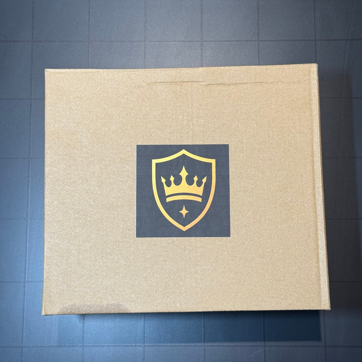 A square brown cardboard box with a dark gray square label in the center featuring a gold crown shield logo, placed on a dark blue-gray tiled surface.