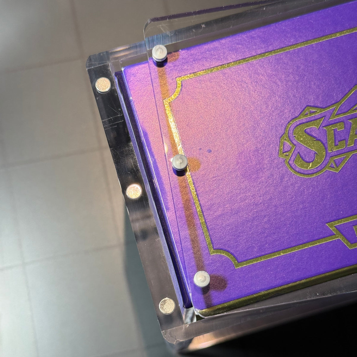 Close-up of a purple rectangular item with a clear acrylic or plastic frame, gold decorative border lines, and a partial gold logo reading 'S.