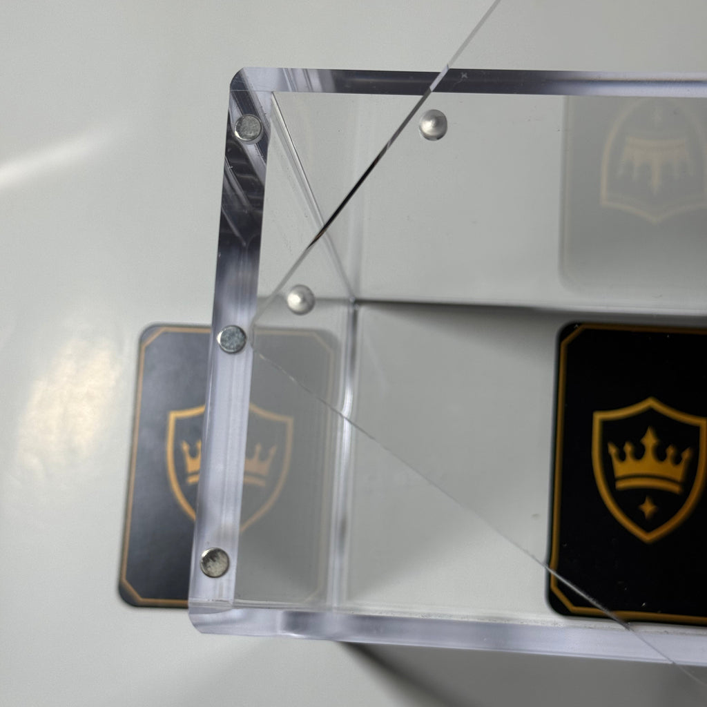 A clear acrylic or glass block with rounded corners and visible metal mounting screws, shown on a light surface with two black cards featuring a gold crown shield logo in the background.